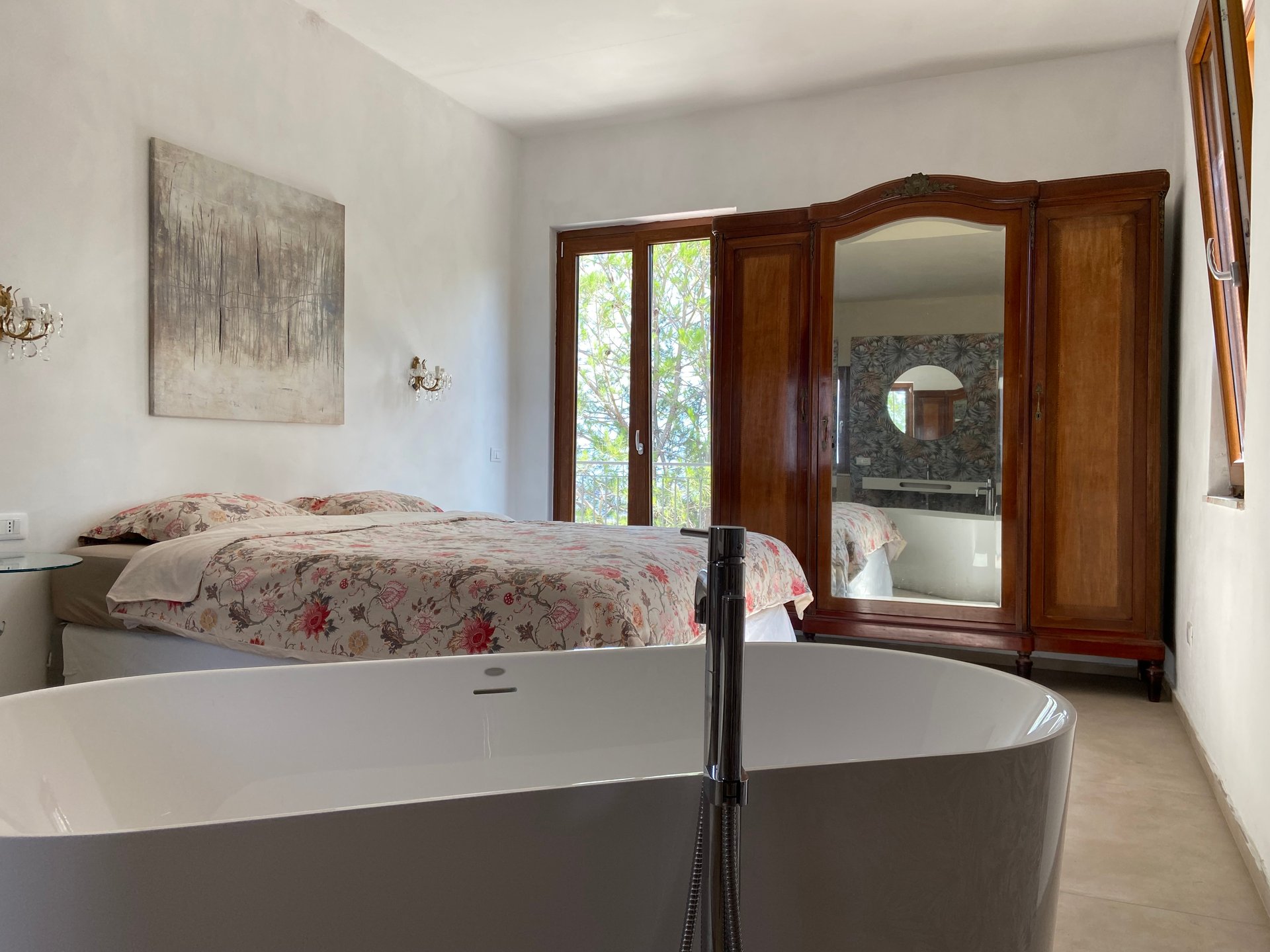 Slaapkamer in Villa Grimiti met een vrijstaand ligbad en een bed met bloemensprei; in de spiegel van de kast is de badkamer te zien